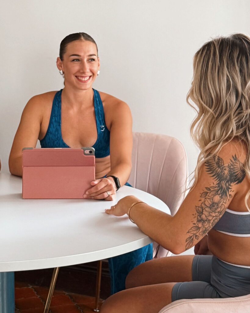 Coaching Fitness : Deux Athlètes Discutent avec une Tablette Deux femmes en tenue de sport discutent autour d'une table blanche ; l'une tient une tablette rose, l'autre a un tatouage floral.