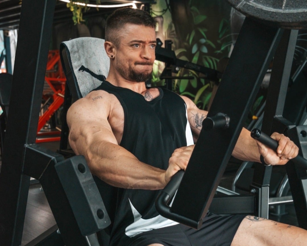 Homme musclé en débardeur noir faisant un exercice de poussée sur une machine de musculation, concentré et en plein effort.