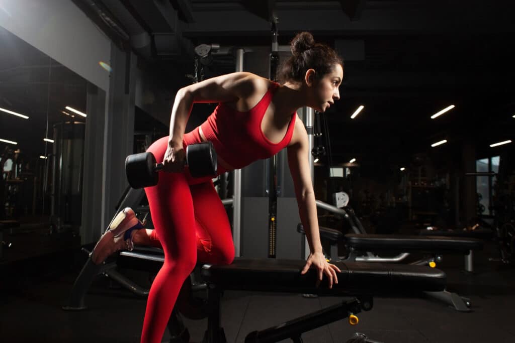 Jeune femme en tenue de sport rouge faisant un rowing haltère sur un banc dans une salle de sport sombre et éclairée.