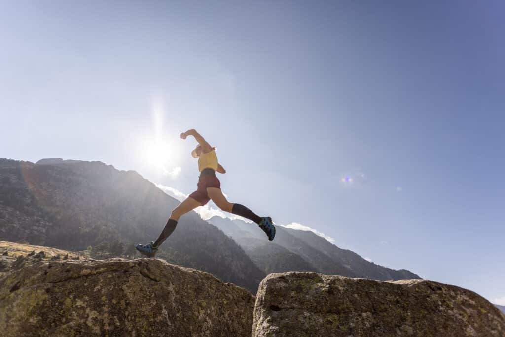 Trail Running Saut Montagne Soleil Action Sport Trail runner en plein saut entre deux rochers, silhouette dynamique face au soleil éclatant et aux montagnes.